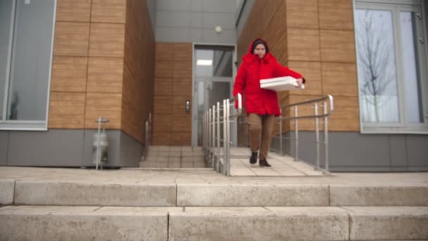 Um mensageiro de roupa vermelha que sai do prédio e tira um pedaço de pizza da caixa de pizza — Vídeo de Stock