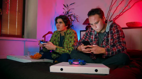Dois jovens sentados na cama um deles jogando o jogo e outro sentado em seu telefone — Fotografia de Stock