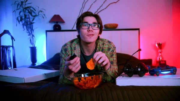 Young interested man in glasses lying on the bed watching TV and eating chips