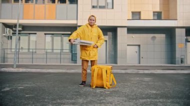 Sarı elbiseli bir kurye elinde pizza kutusuyla yemek dağıtır ve gülümser.