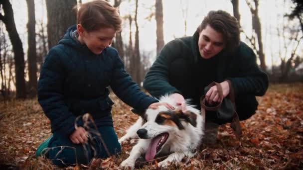 Joven y su hermano pequeño acariciando perro en el parque de otoño — Vídeos de Stock