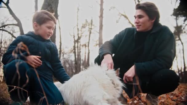 Młody człowiek i jego młodszy brat pieszczą psa w jesiennym parku i rozmawiają — Wideo stockowe