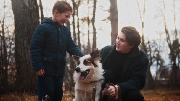 Junger Mann und sein kleiner Bruder streicheln ihren Hund im Herbstpark — Stockvideo