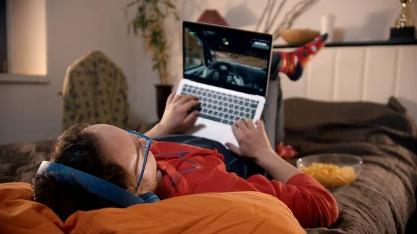 Joven acostado en la cama jugando carrera de videojuegos en el portátil —  Fotos de Stock