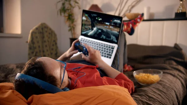 Joven acostado en la cama jugando carrera de videojuegos en el portátil con mando — Foto de Stock