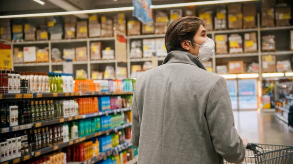 Молодой человек в защитной белой маске ходит между секциями в большом продуктовом магазине — стоковое фото
