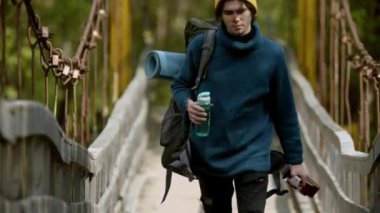 Büyük bir sırt çantası, gitar ve bir şişe su taşıyan genç adam köprüde yürüyor.