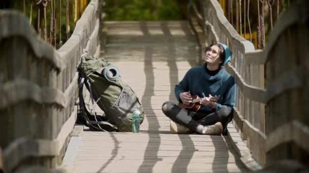 Jovem viajante sentado na ponte e jogando ukulele — Vídeo de Stock