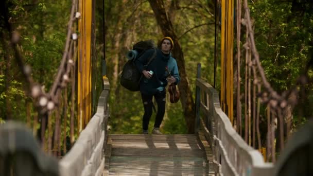 Joven viajero caminando por el puente sosteniendo ukelele y botella de agua — Vídeo de stock