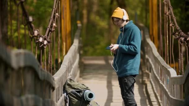 Hombre joven viajero de pie en el puente en los auriculares y tener un chat de vídeo con su amigo — Vídeos de Stock