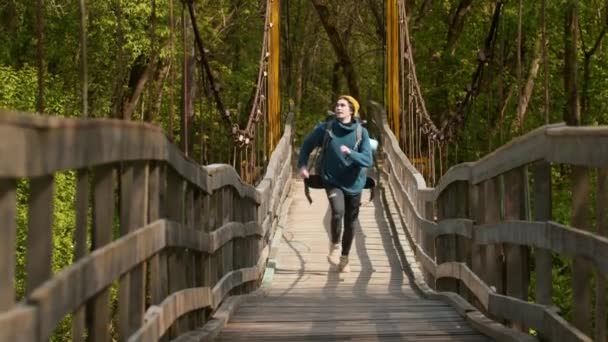 Hombre joven viajero con gran mochila corriendo en el puente en el bosque — Vídeos de Stock