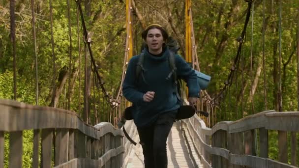 Young man traveler with big backpack running on the bridge in the forest and looking in the camera — Stock Video