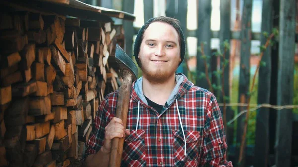 Jovem homem sorridente lenhador com machado de pé na frente da câmera e olhando para a câmera — Fotografia de Stock