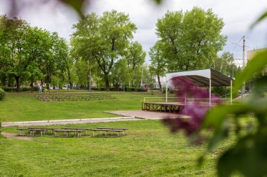 Yaz kenti parkı yağmurdan önce. Yoğun yeşil bitki örtüsü, boş bir sahne ve önünde sıralar..