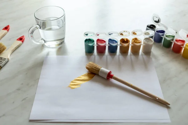 Aydınlık bir masada farklı renkte boya. Çizim için bir kağıt parçası. Fırça darbesi uygulanmış. Fırçalar masanın üstünde ve su temiz bir bardakta.