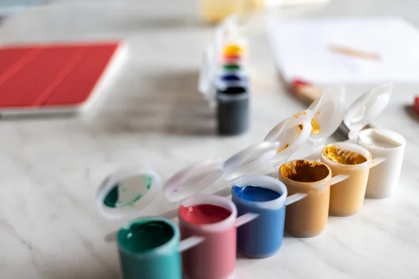 different colors of paint on a light table. on the table is a tablet with a red cover. there is a drawing sheet in the distance. natural light and shadows