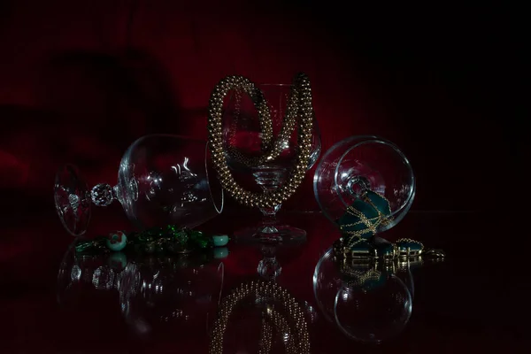 三个项链 其中两个用石头和一个不同寻常的合金 看起来像黄金 用透明的空玻璃杯组成 所有的东西都反映在杯子里 深红色背景 — 图库照片