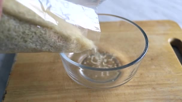 Сырой рис наливают в глубокую прозрачную миску для приготовления или сортировки на деревянной доске. На кухне. дом . — стоковое видео