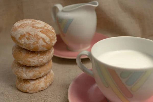milk in a tea cup with pastel strips stands on a light gray background. near the milk container. Both on a pink plates. gingerbread or cookies are stacked on top of each other.