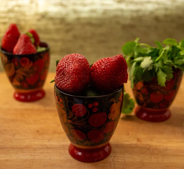 a composition of three painted cups with berries in the drawing and strawberries inside. in the third glass of greens. they stand on a wooden cutting Board. light grey background