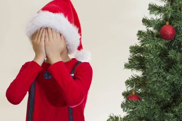 Niño en gorra Santa roja escondiendo su cara con ambas manos cerca del árbol de navidad — Foto de Stock