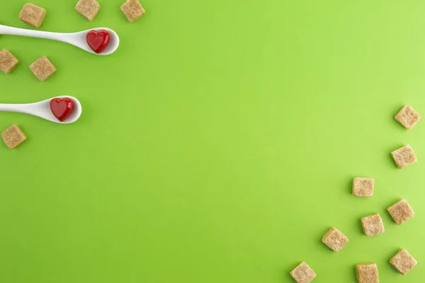 Two white spoons with red hearts on them and brown sugar cubes over greenery background. Top view. Copy space for text. — Stock Photo, Image