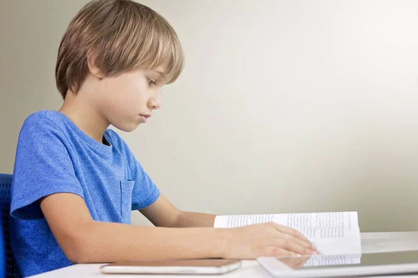 Geconcentreerde kind lezen van een boek. Mobiele telefoon en tablet pc zijn op de tafel in de buurt van hem. Technologie, onderwijs, school concept — Stockfoto