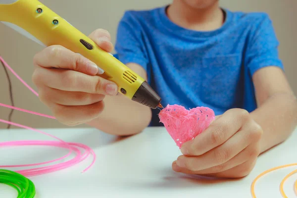 Crianças mão segurando caneta de impressão 3D amarelo com filamentos e faz o coração no fundo branco. Vista superior. Copiar espaço para texto. Foco seletivo — Fotografia de Stock