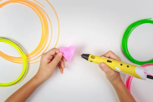 La mano de los niños sostiene la pluma de impresión 3D amarilla con filamentos y hace que el corazón sobre fondo blanco. Vista superior. Copia espacio para texto. Enfoque selectivo — Foto de Stock