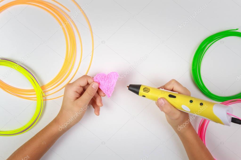 Kids hand holding yellow 3D printing pen with filaments and makes heart on white background. Top view. Copy space for text. Selective focus