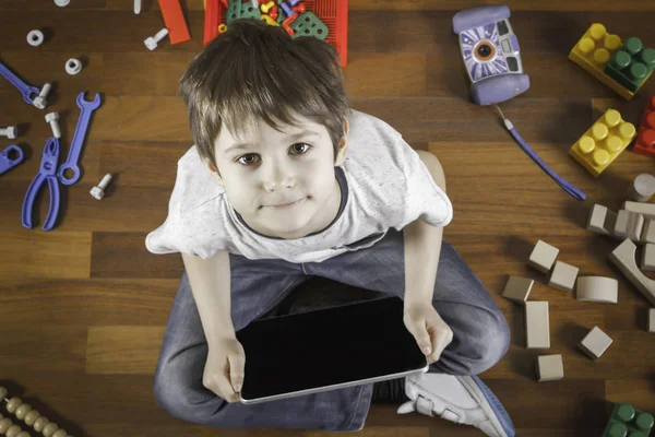 Rapaz a brincar com o computador tablet. Brinquedos coloridos ao redor dele no chão de madeira. Vista superior — Fotografia de Stock