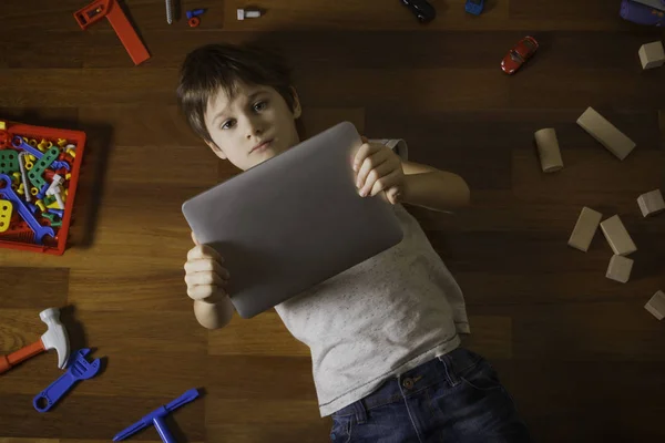 Rapaz a brincar com o computador tablet. Criança deitada no chão, muitos brinquedos ao seu redor no chão de madeira . — Fotografia de Stock