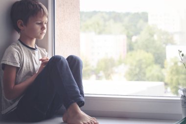 Pencere kalkan üzerinde oturan ve pencereden dışarı bakarak üzgün çocuk