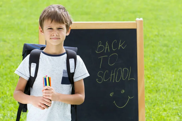 Liten skolpojke med pennor och ryggsäck mot tavlan. Utbildning, tillbaka till skolan-konceptet — Stockfoto
