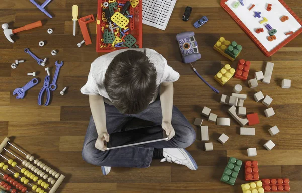 Rapaz a brincar com o computador tablet. Muitos brinquedos à volta dele no chão de madeira. Vista superior — Fotografia de Stock