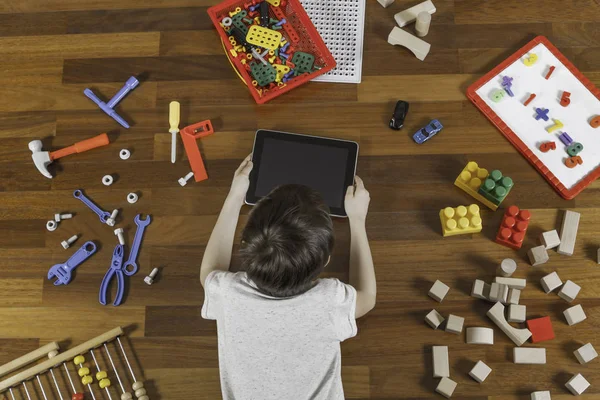 Um miúdo a mentir e a brincar com o computador tablet. Muitos brinquedos à volta dele no chão de madeira. Vista superior — Fotografia de Stock