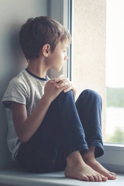 Pencere kalkan üzerinde oturan ve pencereden dışarı bakarak üzgün çocuk