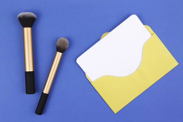 Maquillaje cepillos y sobre amarillo con tarjeta en blanco sobre fondo azul — Foto de Stock