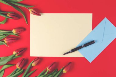 Boş kart, kalem, mavi bir zarf içerisinde ve kırmızı bahar lale çiçekleri kırmızı zemin üzerine.