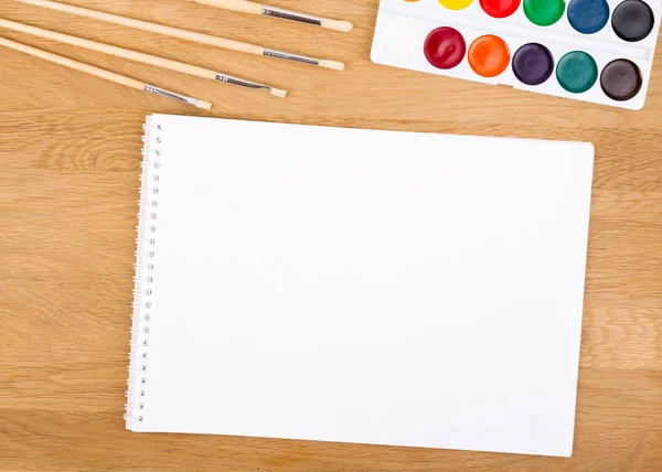 Watercolor paints, brushes and blank white paper sheet on wooden background.