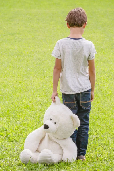 Rapaz triste e solitário com ursinho de peluche no prado. Criança a olhar para baixo. Vista traseira. Tristeza, medo, conceito de solidão — Fotografia de Stock