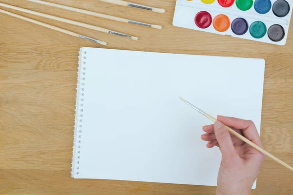 Vista superior de una mano con pincel de pintura, acuarelas y hoja de papel en blanco —  Fotos de Stock
