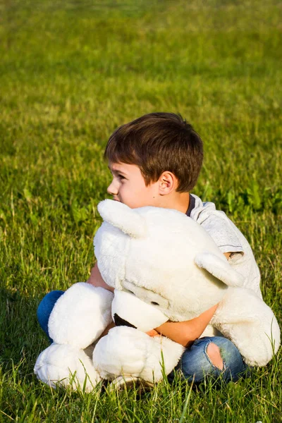 Pequeño niño abrazando oso de peluche mientras está sentado en la hierba . — Foto de Stock