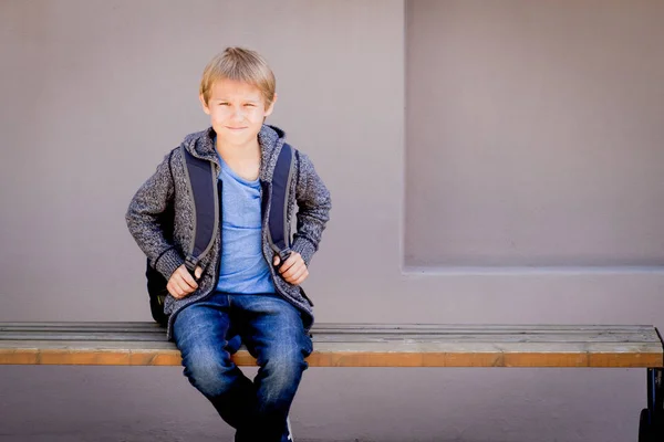 Pojke med ryggsäck som sitter på bänken nära skolan — Stockfoto