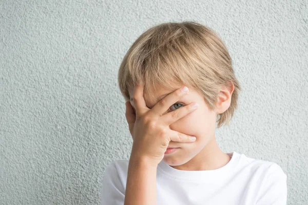 Yüzünü eli ile kapsayan ve gri arka plan karşı parmak kameradan bakarak çocuğun