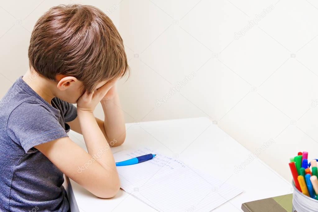 Little kid made his homework at home. The boy is tired and covers his face with hands