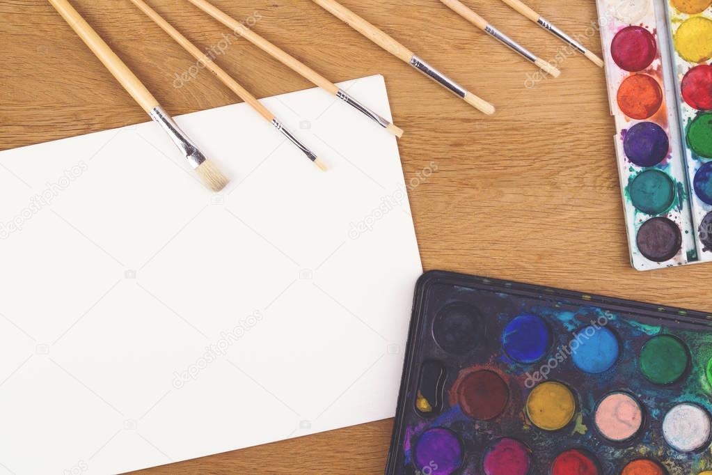 Watercolor paints, brushes for painting and blank white paper sheet on wooden background.