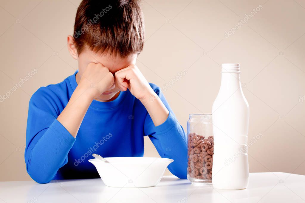 Sleepy kid sitting at the table early in the morning. Boy do not want to eat his breakfast