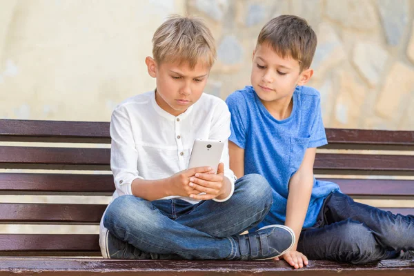 Niños con teléfono móvil sentados en el banco al aire libre — Foto de Stock