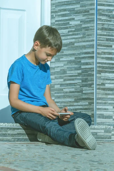Menino com telefone celular sentado na escada em frente a uma porta — Fotografia de Stock